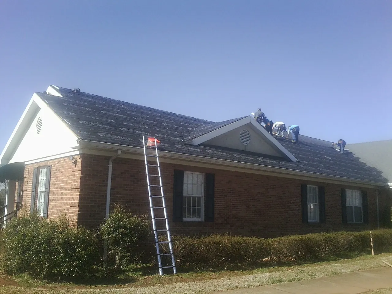 Crew tearing shingles off a brick ranch roof with a ladder set — Metal Roof Repair work by our crew in Central Square