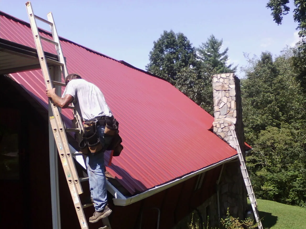 Roofer on a ladder beside a red metal roof and stone chimney — Soffit & Fascia Repair by Roofing Brothers in Central Square
